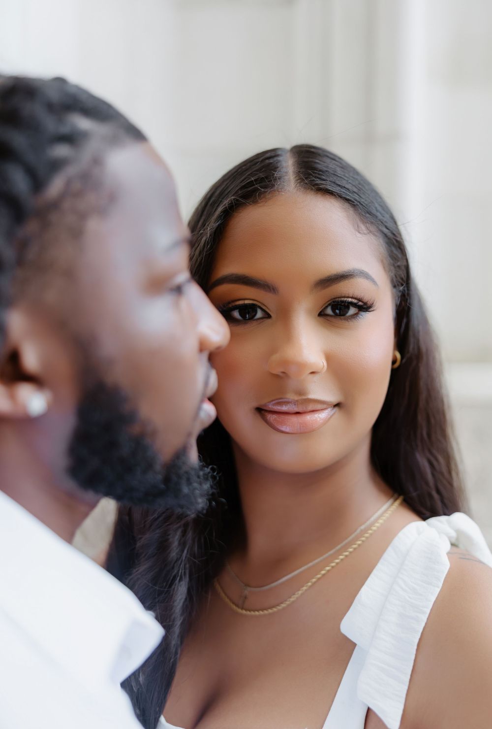 Close-up of a couple sharing an intimate moment, woman in white dress with gold necklace smiling softly.