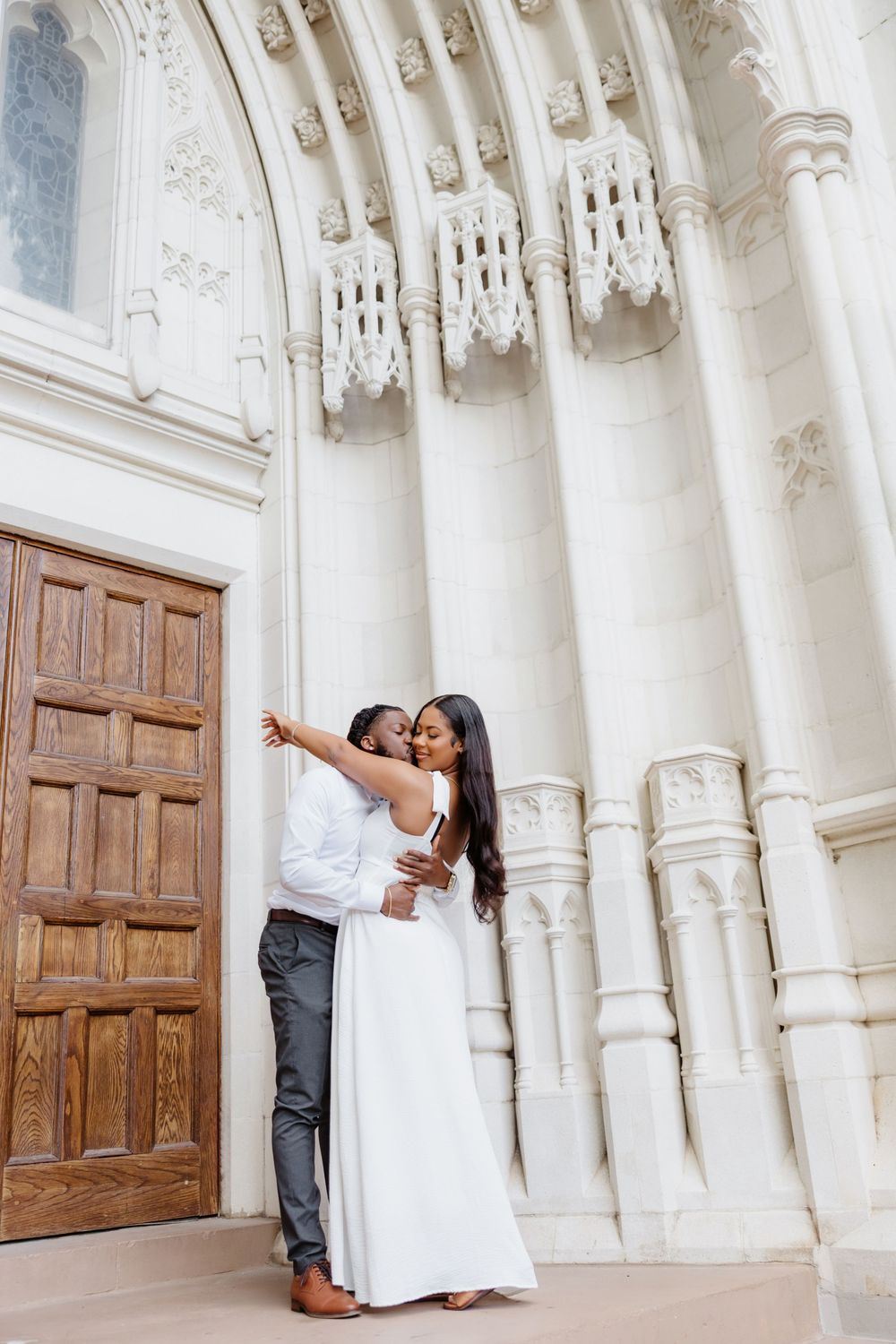 Couple embracing and kissing in front of ornate white stone church entrance with wooden door.
