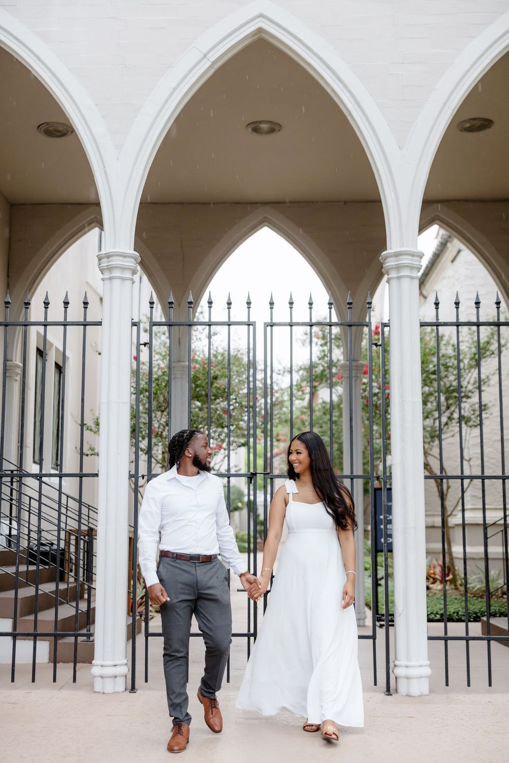Couple holding hands smiling at each other under white Gothic arches with iron gate backdrop.