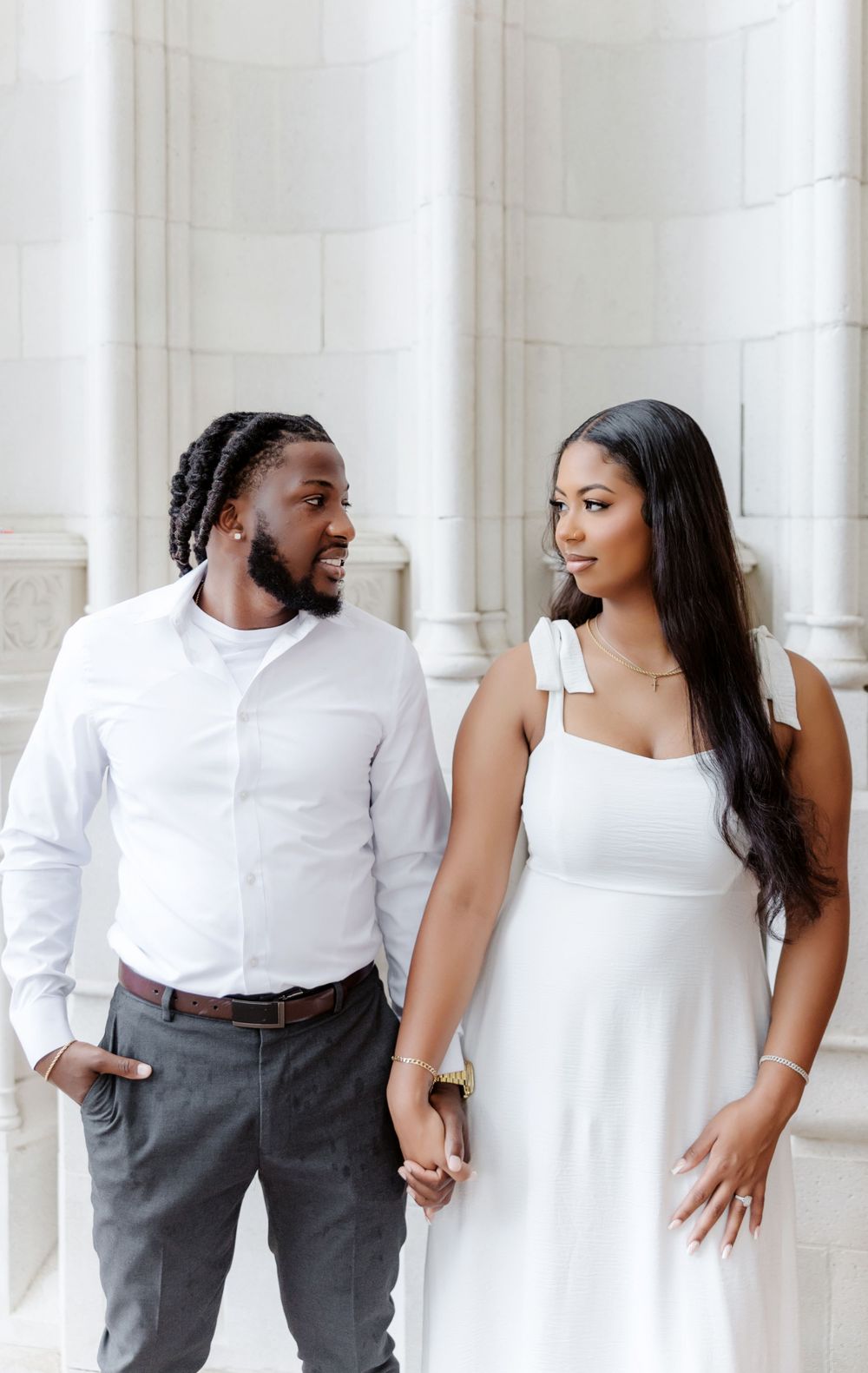 Elegant couple holding hands in white outfits before classic white columns in a romantic engagement photo.