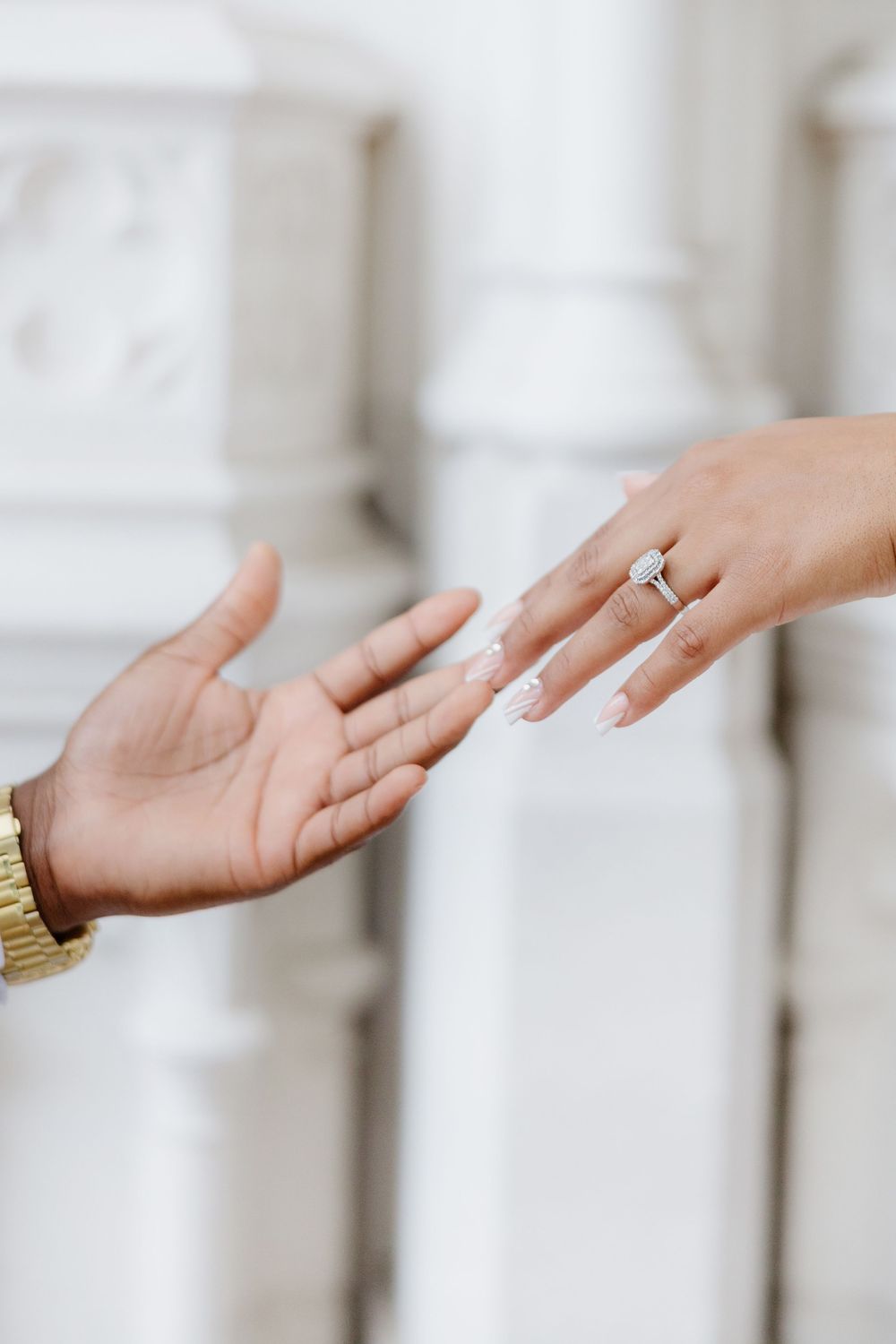 Two hands reaching toward each other, one wearing a diamond engagement ring with elegant white nail art. Bride-to-be showing her engagement