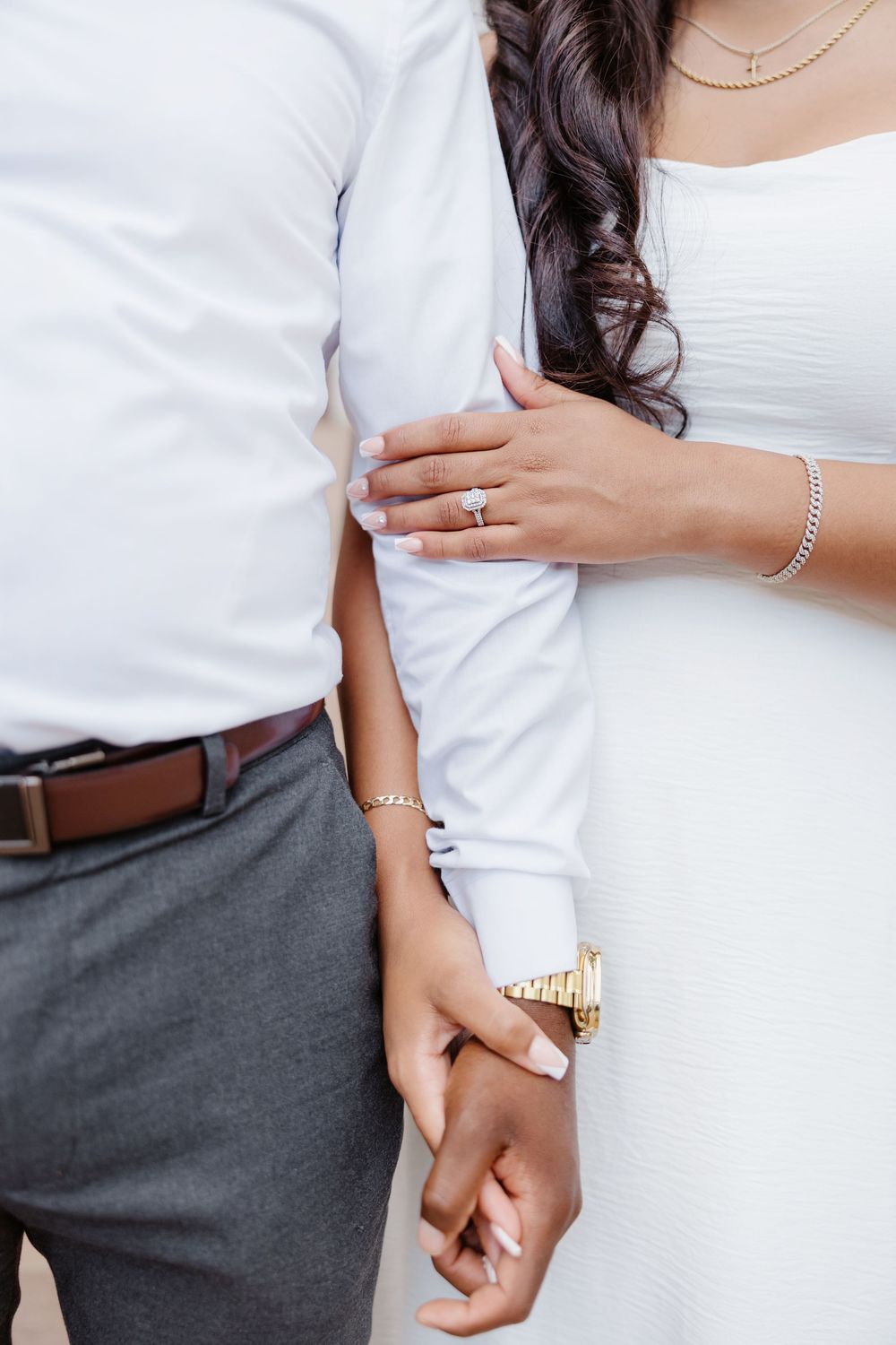 Couple holding hands, woman wearing engagement ring, bracelet, and gold watch, man in white shirt and gray pants.