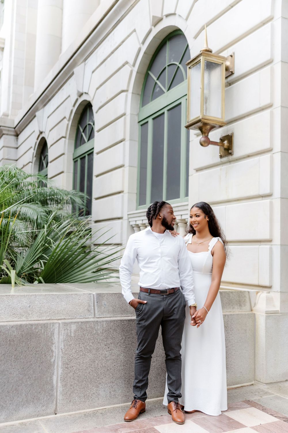 A couple in white outfits holds hands and smiles outside an elegant building with arched windows.