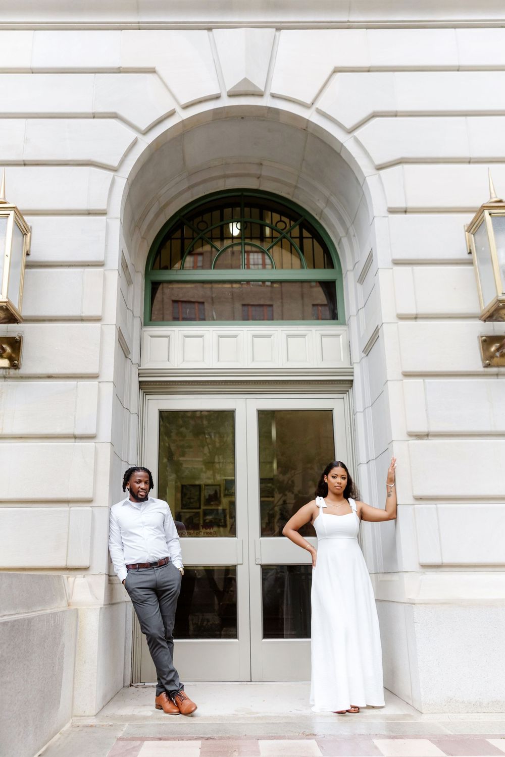 Elegant couple in white attire poses before grand arched doorway of classic white stone building.