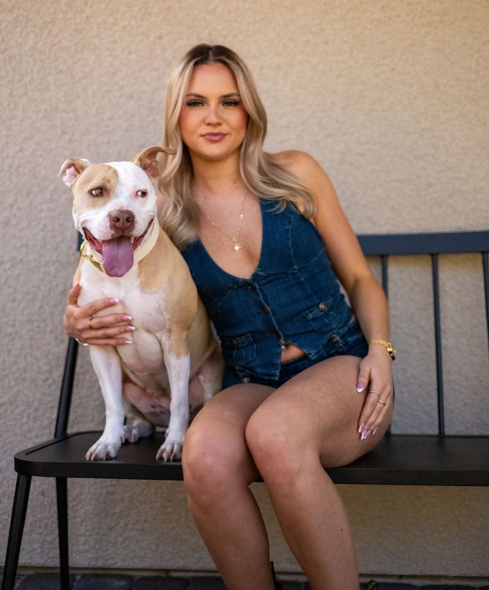 Blonde woman in denim romper sitting on bench with white and brown pit bull dog outdoors.
