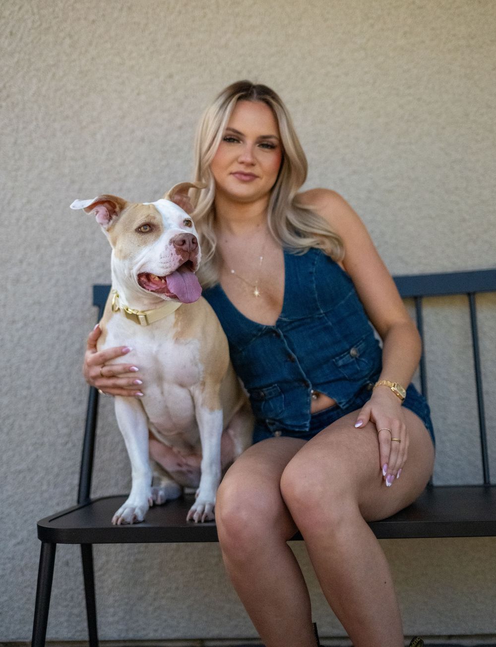 Blonde woman in denim outfit sitting on bench with white and brown pit bull dog outdoors.
