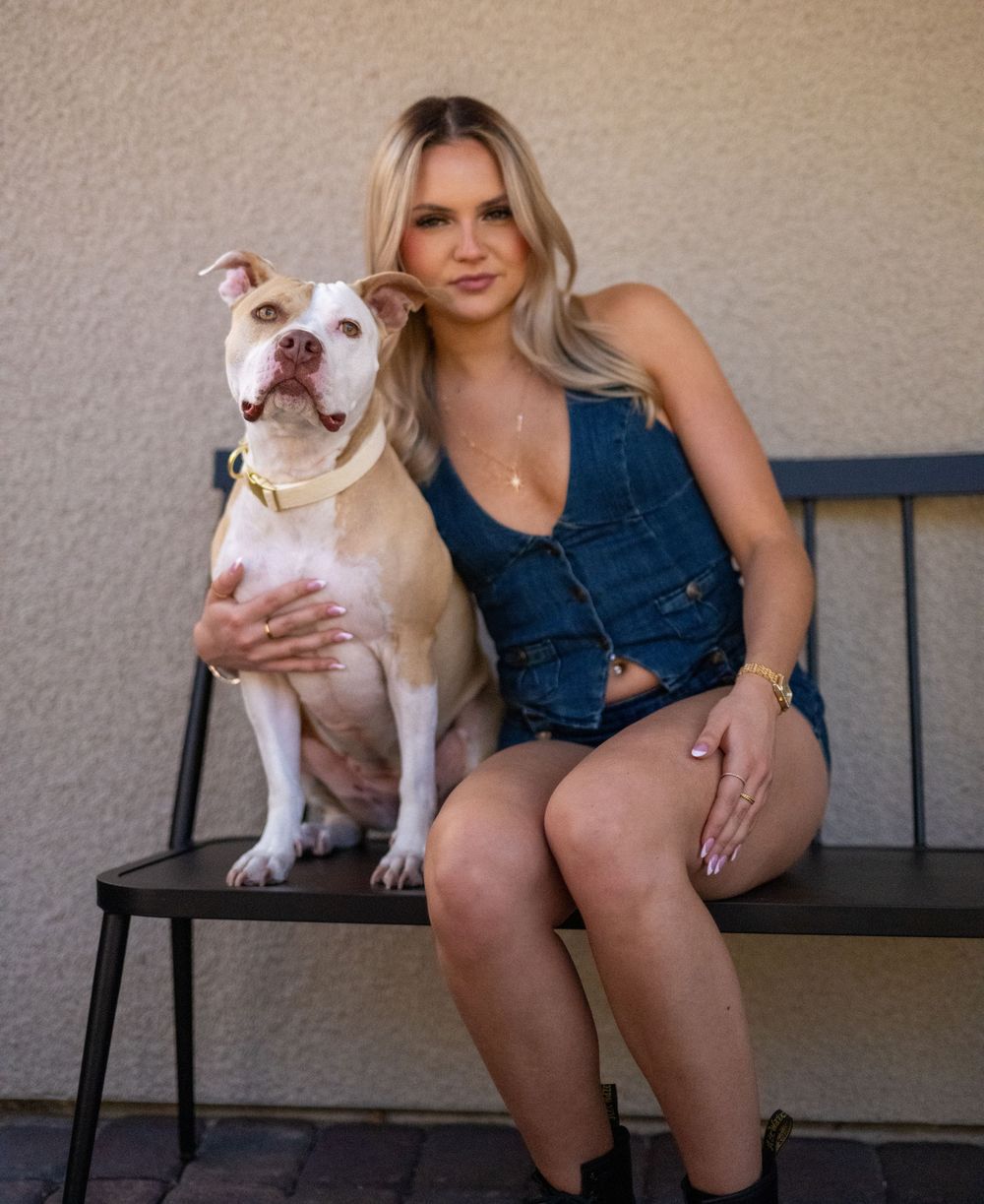 Blonde woman in denim outfit sitting on bench with white pit bull dog outdoors.