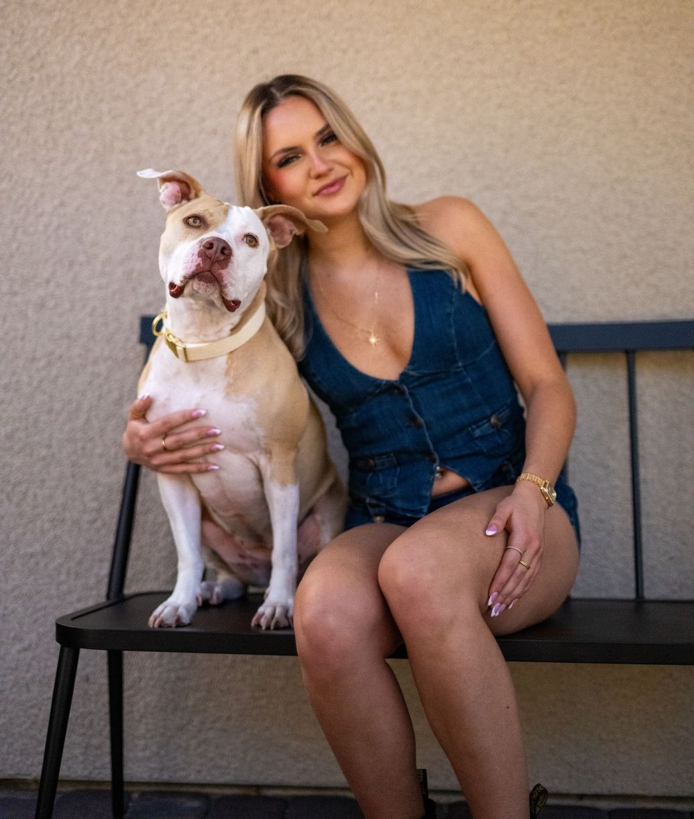 Smiling blonde woman in blue denim top posing with white pit bull on outdoor bench.