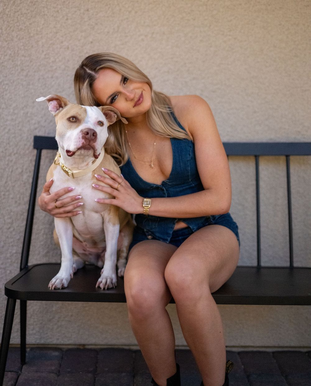 Blonde woman in denim outfit sitting on bench hugging white pit bull dog with gold collar.