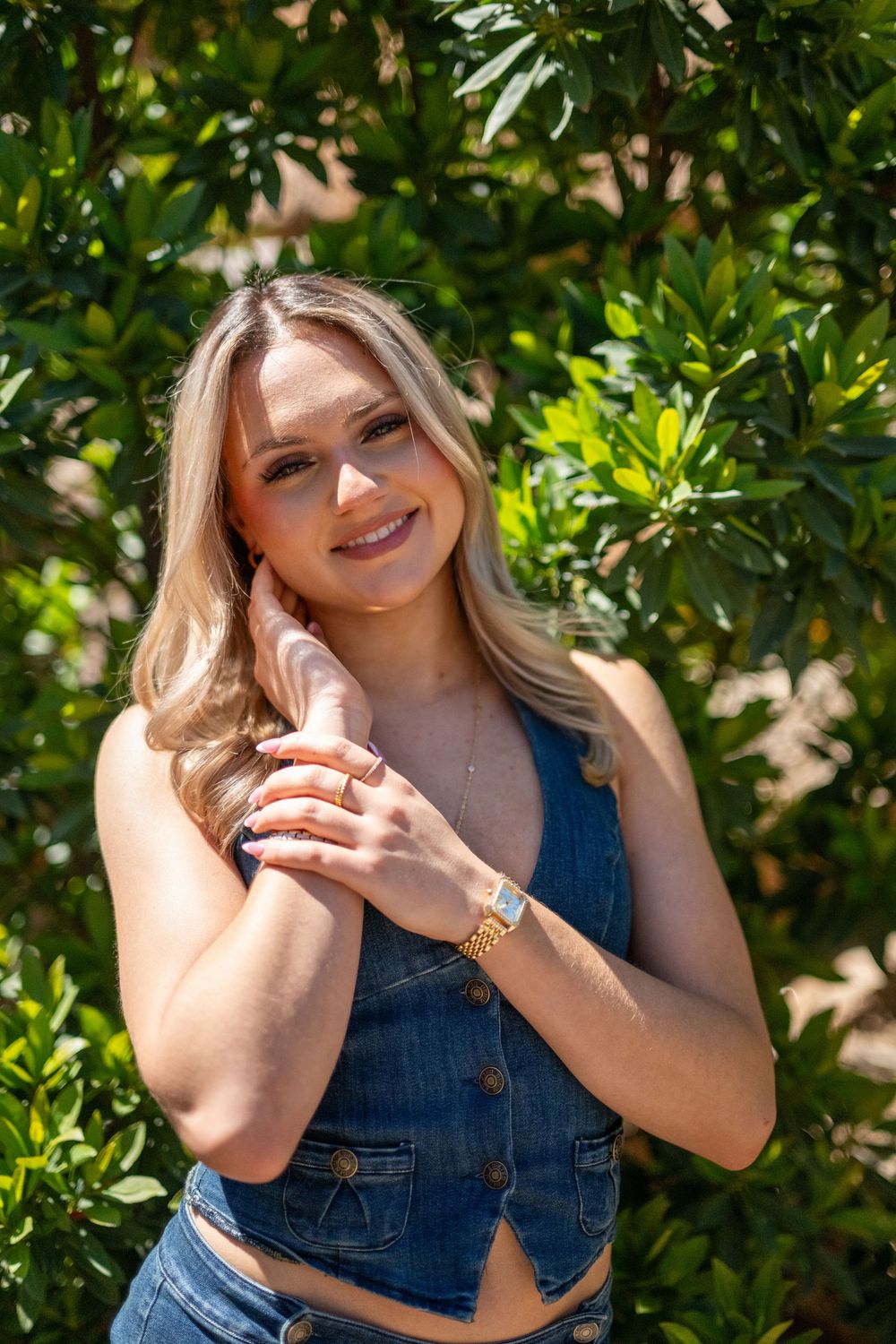 Smiling blonde woman in denim vest top posing outdoors among lush green bushes on a sunny day.