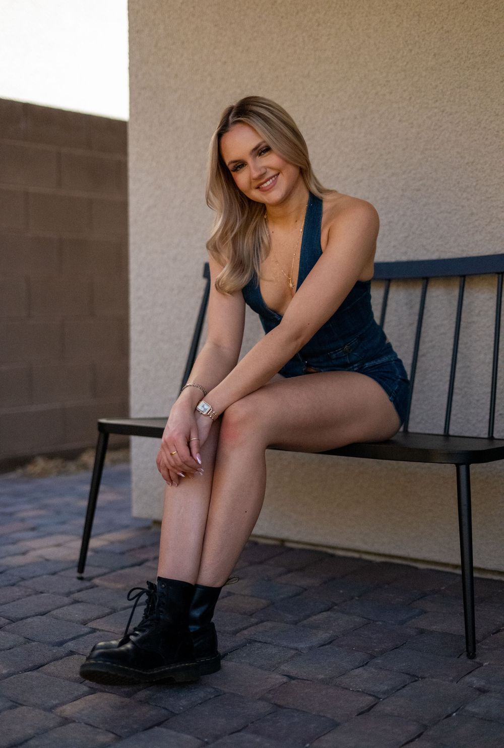 Smiling blonde woman in teal romper and black boots sitting on a bench outdoors.