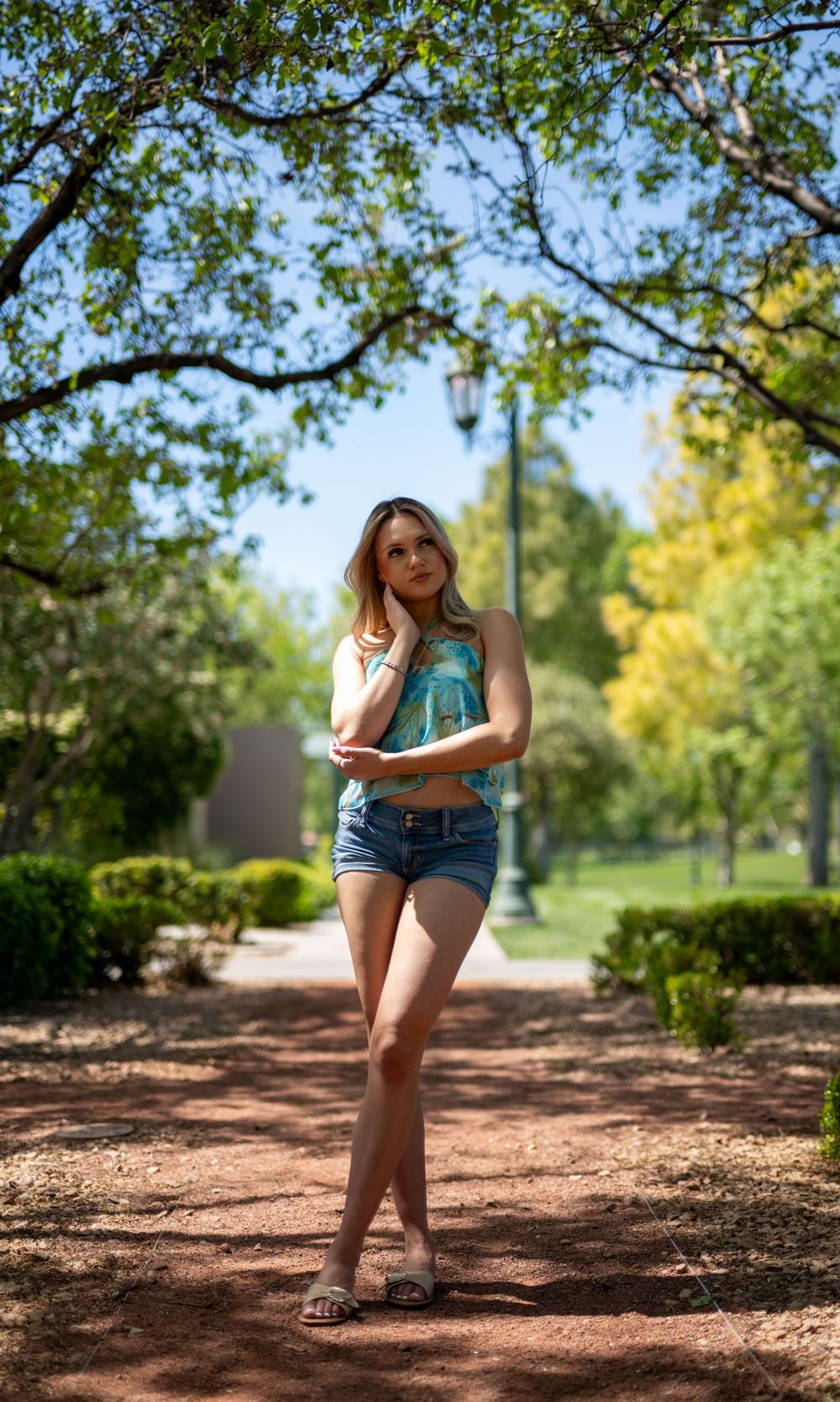 Blonde woman in floral top and denim shorts posing on a tree-lined park path in sunlight.