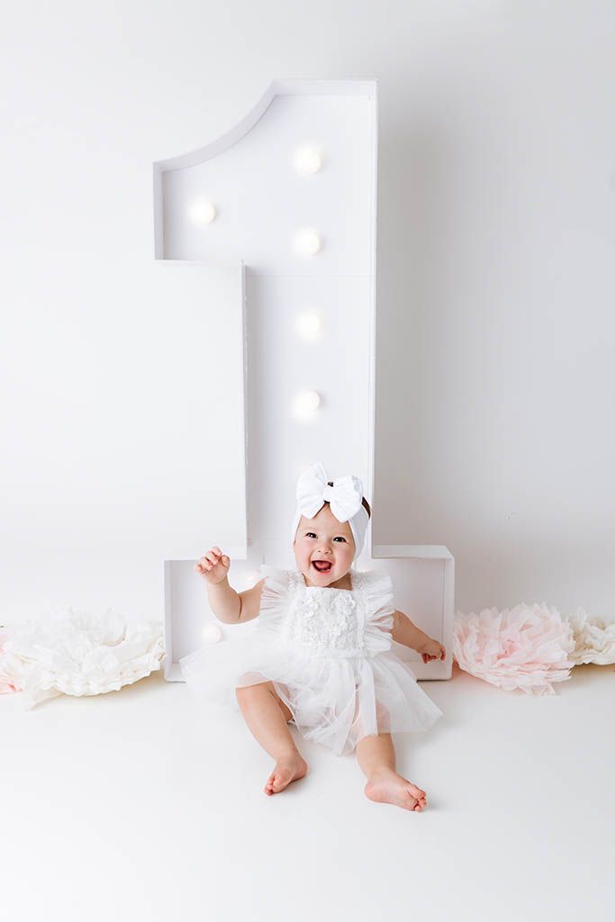A joyful baby girl in a white dress celebrates her first birthday beside a large illuminated number one decoration.