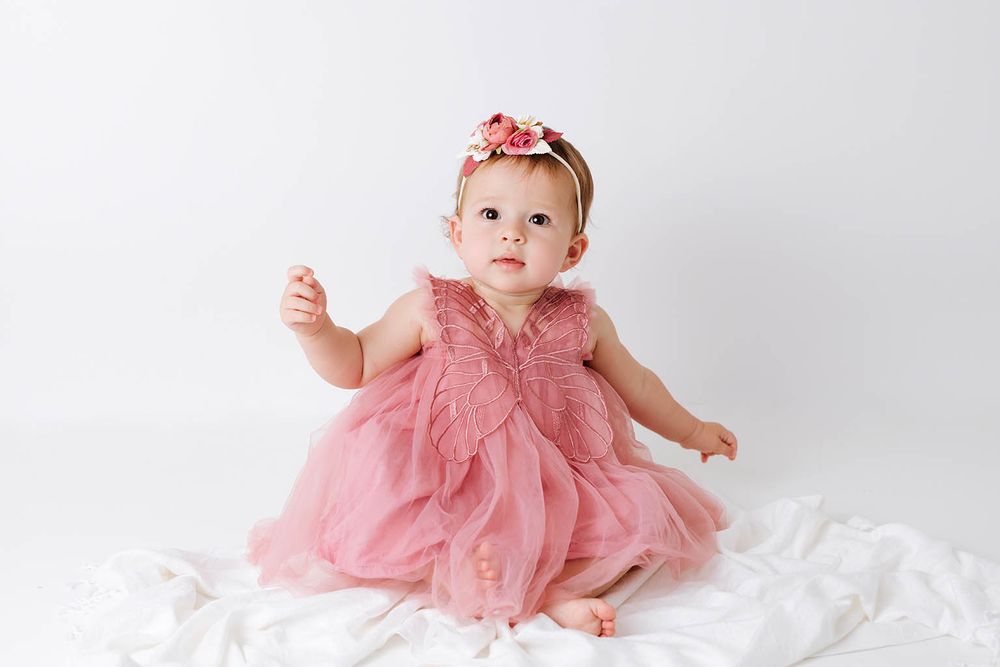 Adorable baby girl in pink ruffle tulle dress with floral headband sitting on white fabric backdrop.