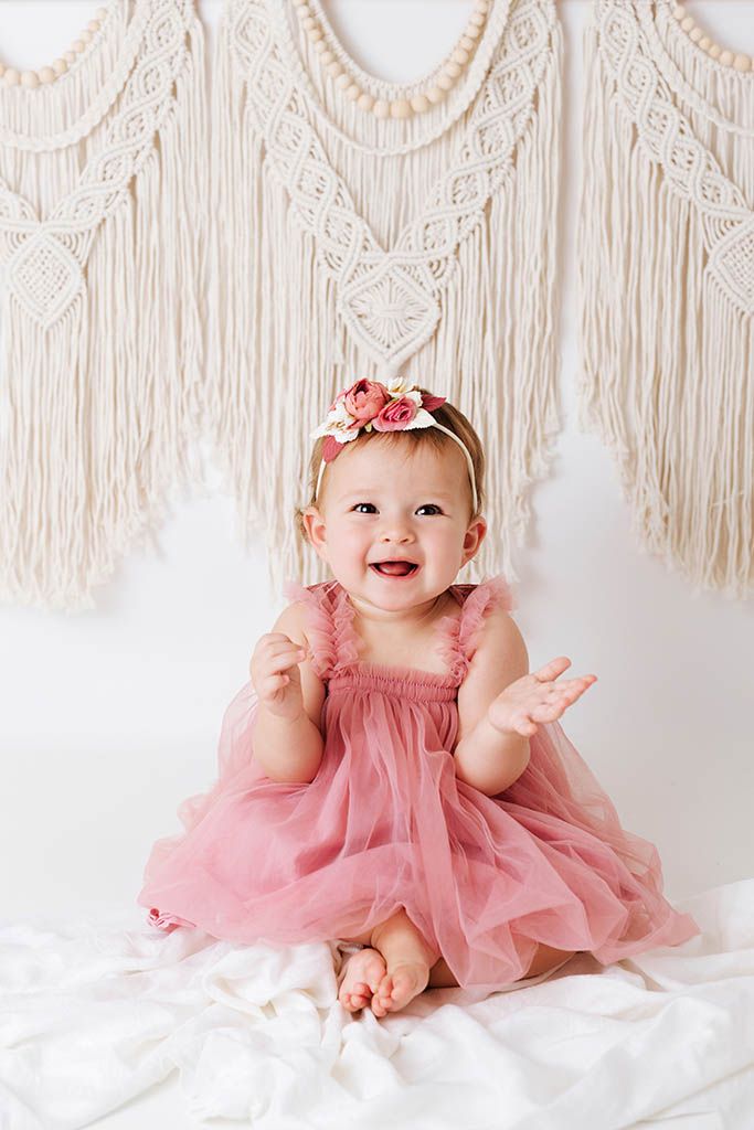 Happy baby girl in pink tulle dress with floral headband sitting before boho macrame wall hanging.