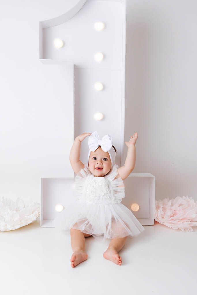 A baby girl in a white tutu dress and bow headband sits in front of a large illuminated number one on her first birthday.