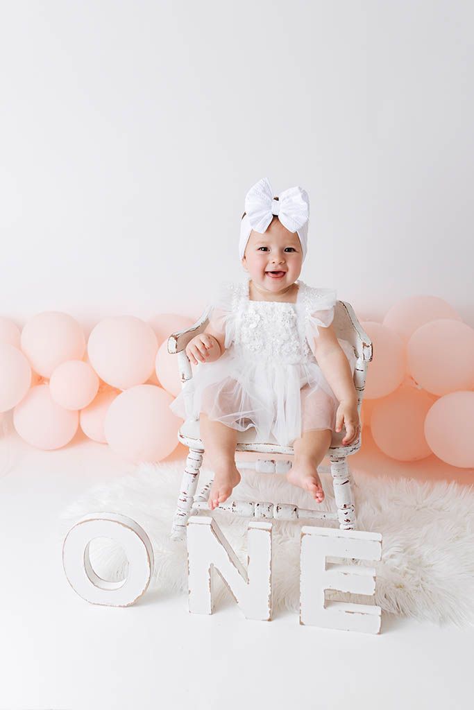 Baby girl in white dress and bow headband sits on chair with ONE letters and pink balloons for first birthday photo.