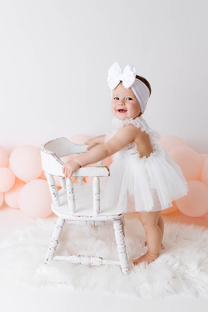 Smiling baby girl in white tutu dress and bow headband standing by vintage chair with pink balloons.