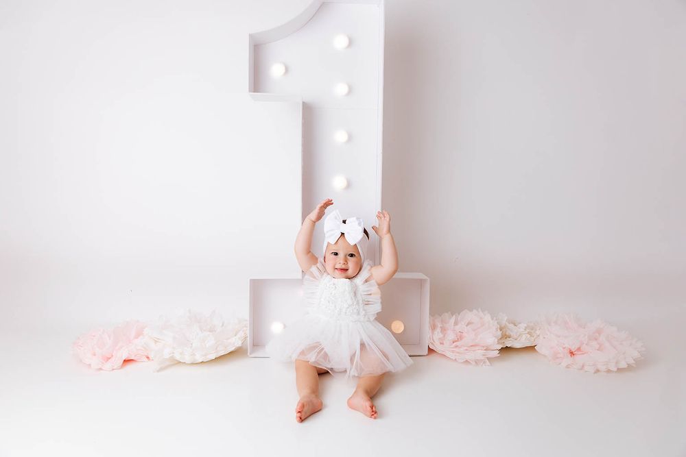 Baby girl in white tutu dress celebrates first birthday beside illuminated number one with pink floral decorations.