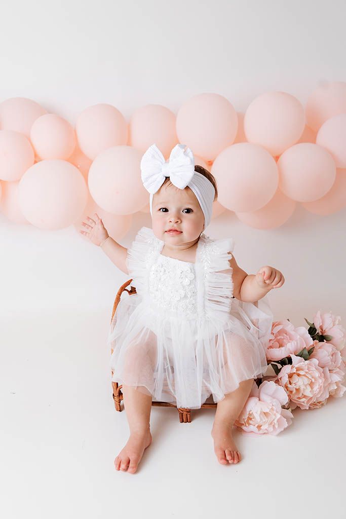Baby girl in white tulle dress with bow headband sits in chair surrounded by pink balloons and peonies.