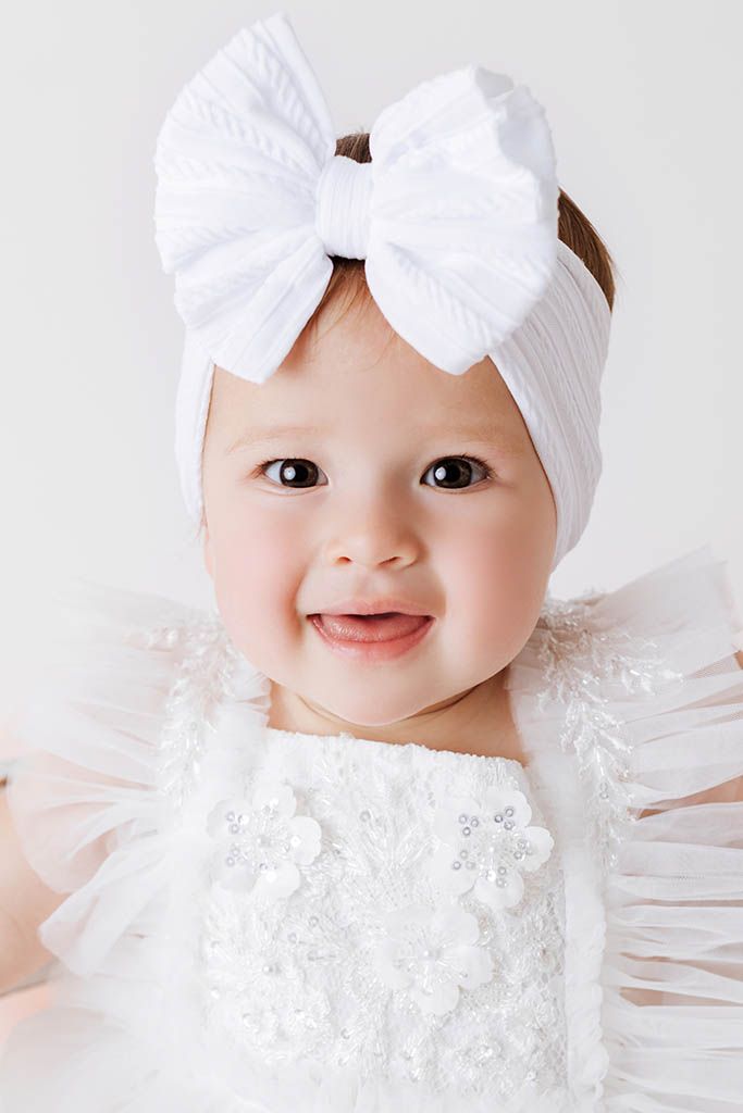 Smiling baby girl in white lace dress and large white bow headband poses for a portrait.