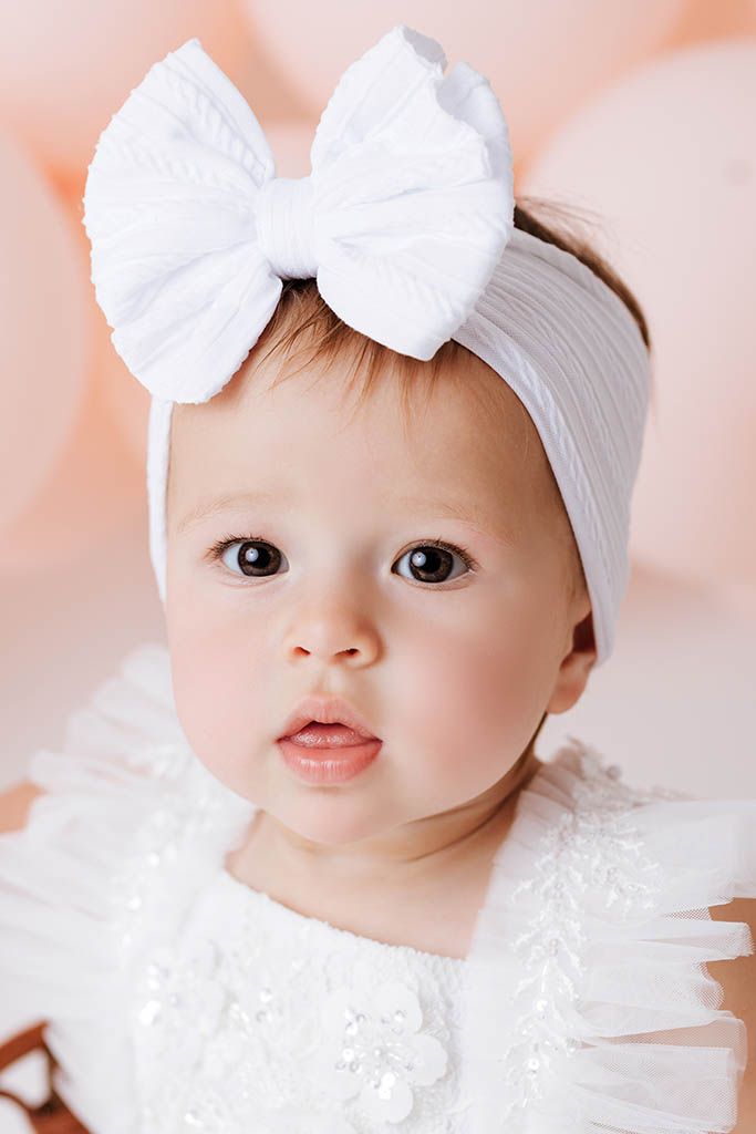 Close-up portrait of adorable baby girl wearing white bow headband and white lace dress.