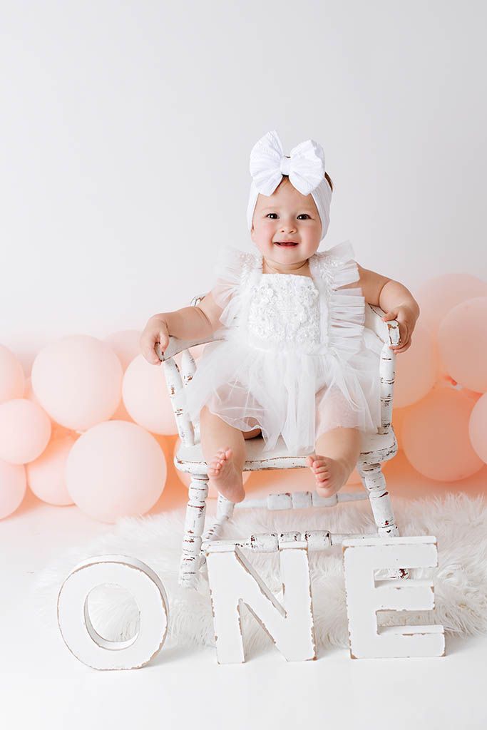 Baby girl in white dress and bow headband sits on chair with ONE letters and pink balloons for first birthday.