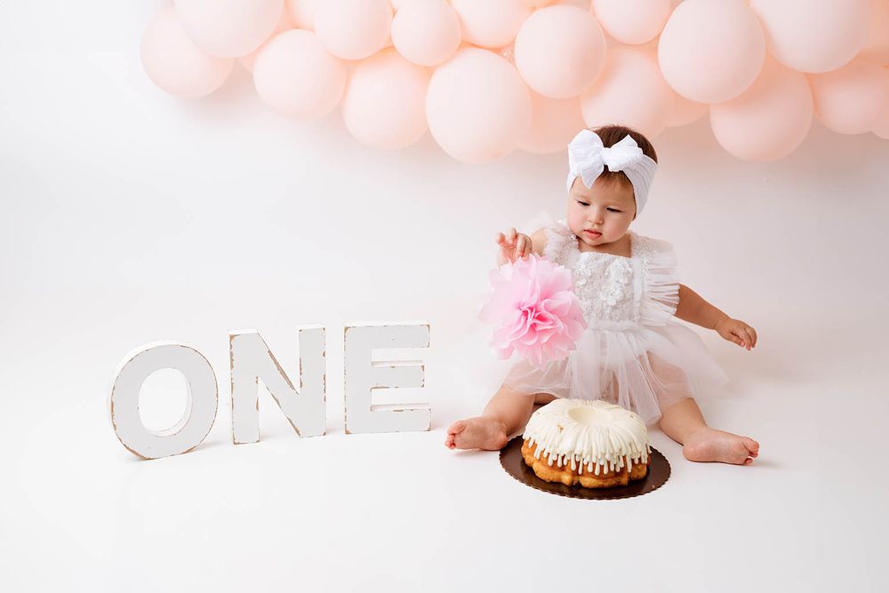 Baby girl in white tutu dress sits beside ONE sign with pink cake and balloon arch for first birthday cake smash.