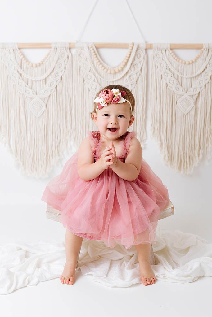 Happy toddler girl in pink tulle dress with floral headband stands before boho macrame backdrop.