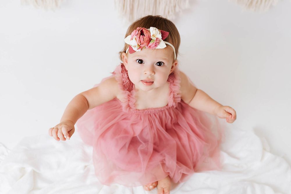 Cute baby girl in dusty rose tulle romper with floral headband sitting on white blanket.