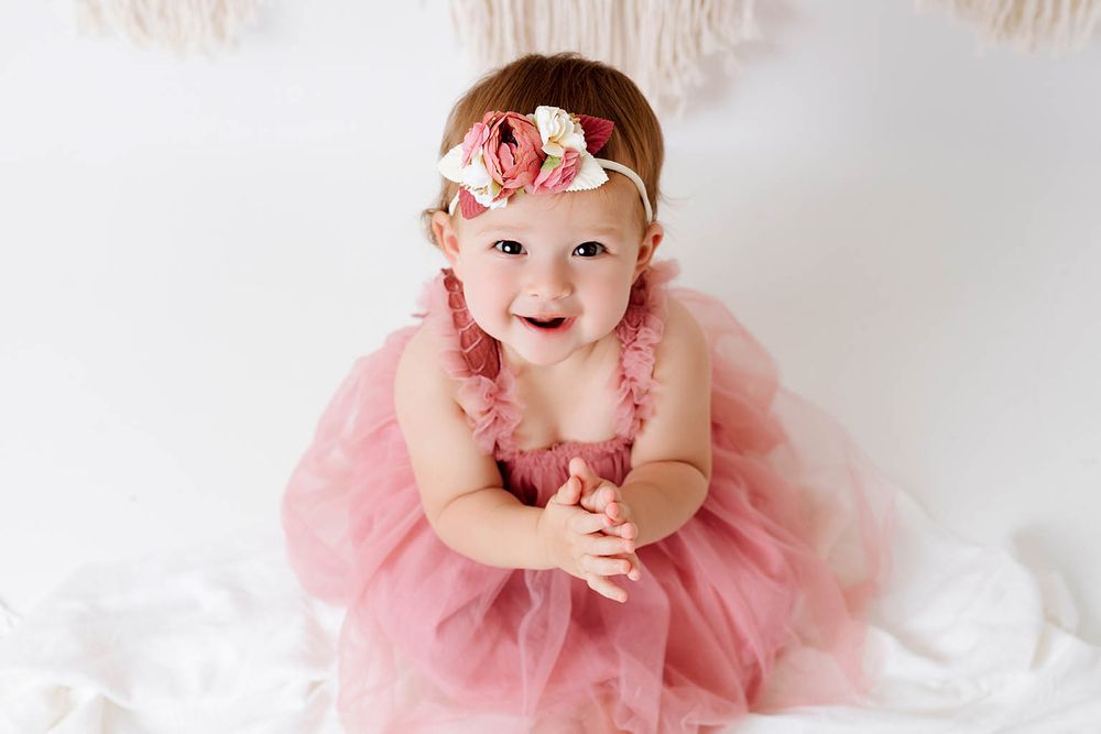 Adorable smiling baby girl in pink tutu dress with floral headband poses for first birthday portrait photo.