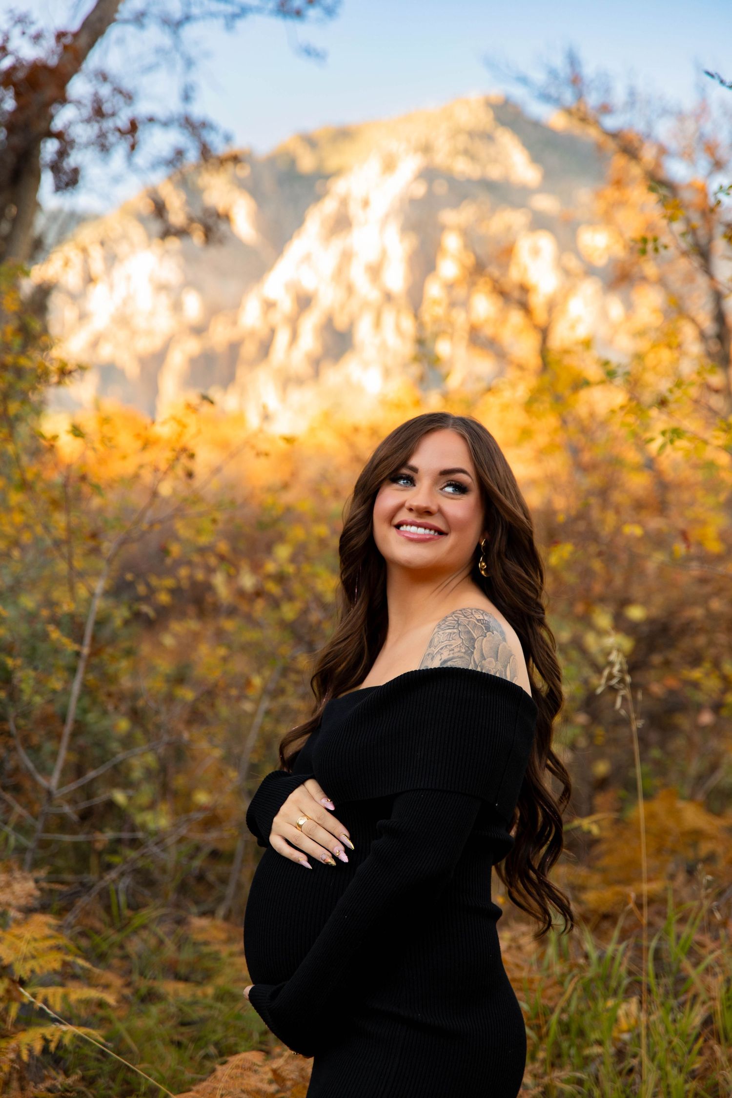 Expecting mother in black dress poses outdoors against mountain backdrop with autumn foliage.