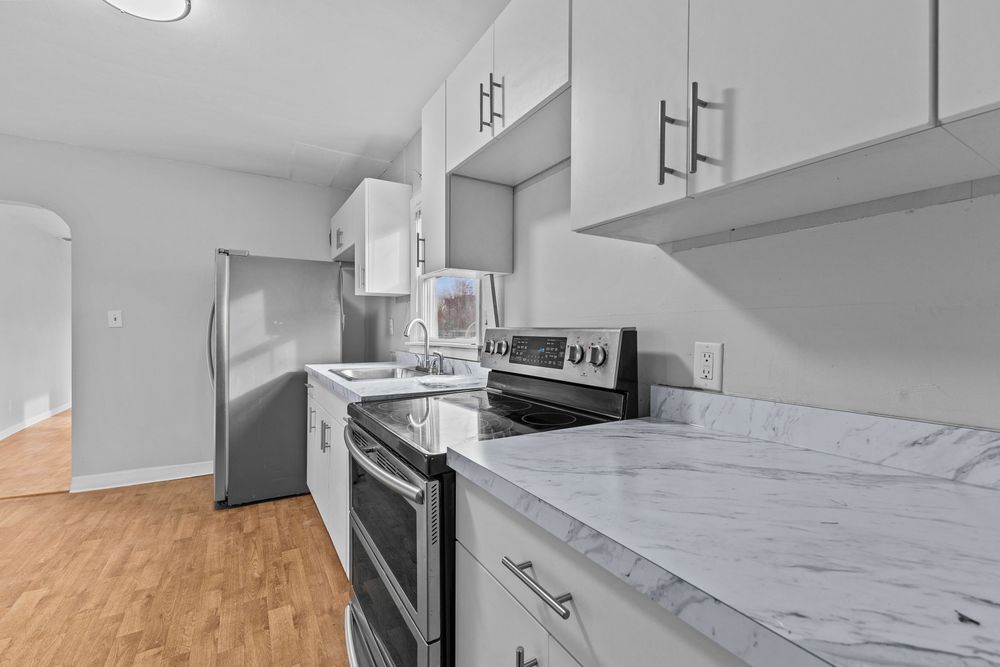 Galley-style kitchen with white cabinets, marble countertops, stainless steel appliances, and hardwood floors.