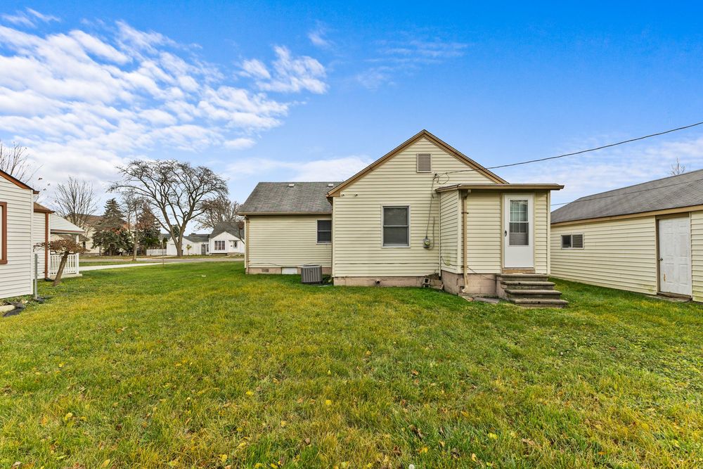 Rear exterior of yellow bungalow with back door entry, large green lawn, and neighboring homes visible.