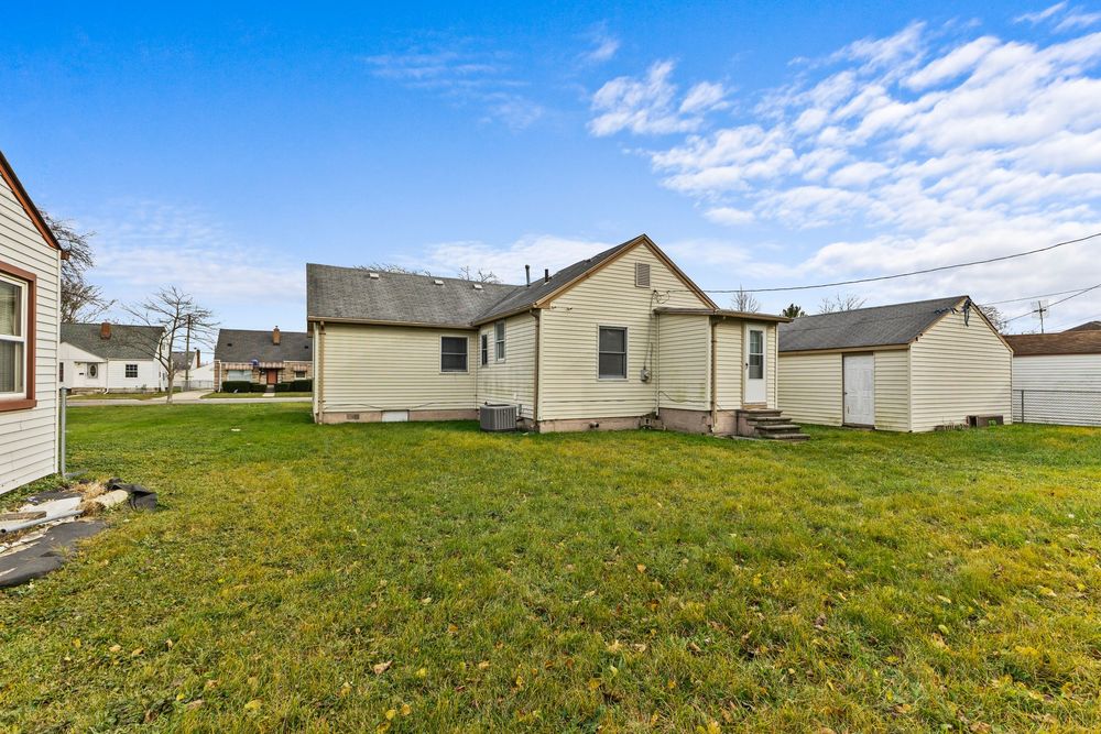 Rear exterior of a bungalow home with green lawn, small deck, detached garage, and neighboring houses.