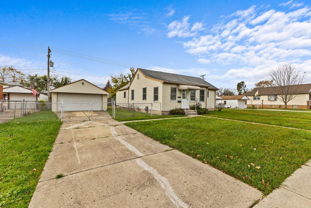 Charming yellow bungalow with detached garage, concrete driveway, and well-maintained lawn under a blue sky.