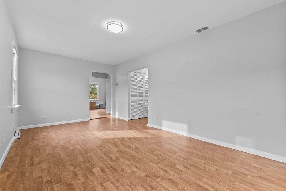 Bright empty living room with light hardwood floors, white walls, and doorway leading to adjacent room.