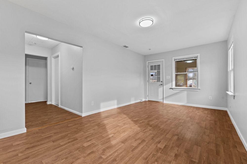 Spacious empty room with medium-tone hardwood floors, white walls, and window providing natural light.
