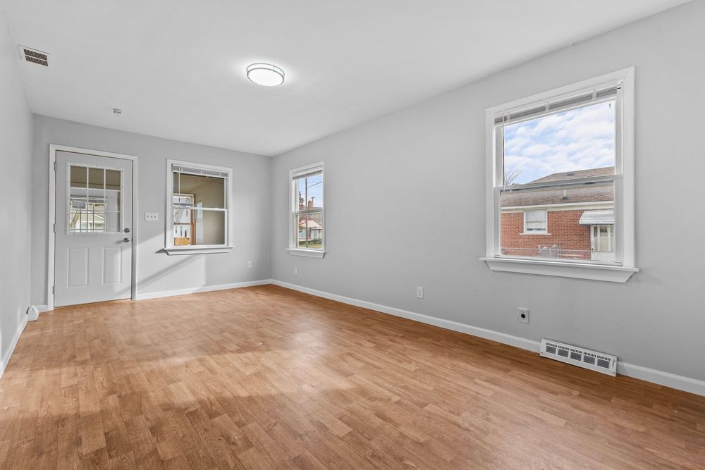 Sun-filled empty living room with hardwood floors, multiple windows, and front entry door.
