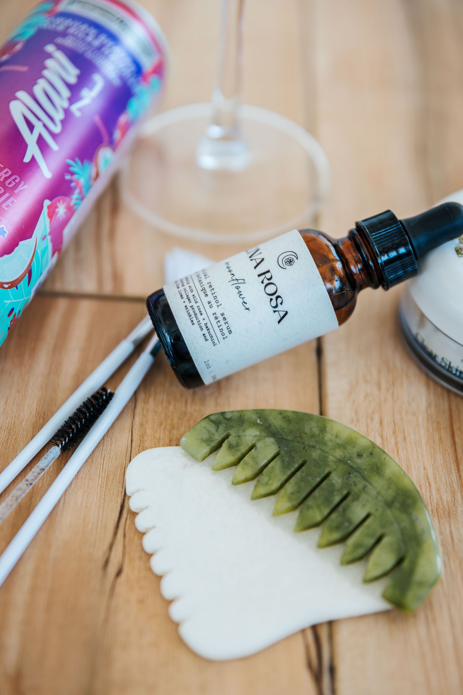 Gua sha tools, facial serum, spoolie brushes, Alani Nu can, and wine glass on wooden surface. Spa and facial skincare branding pictures.