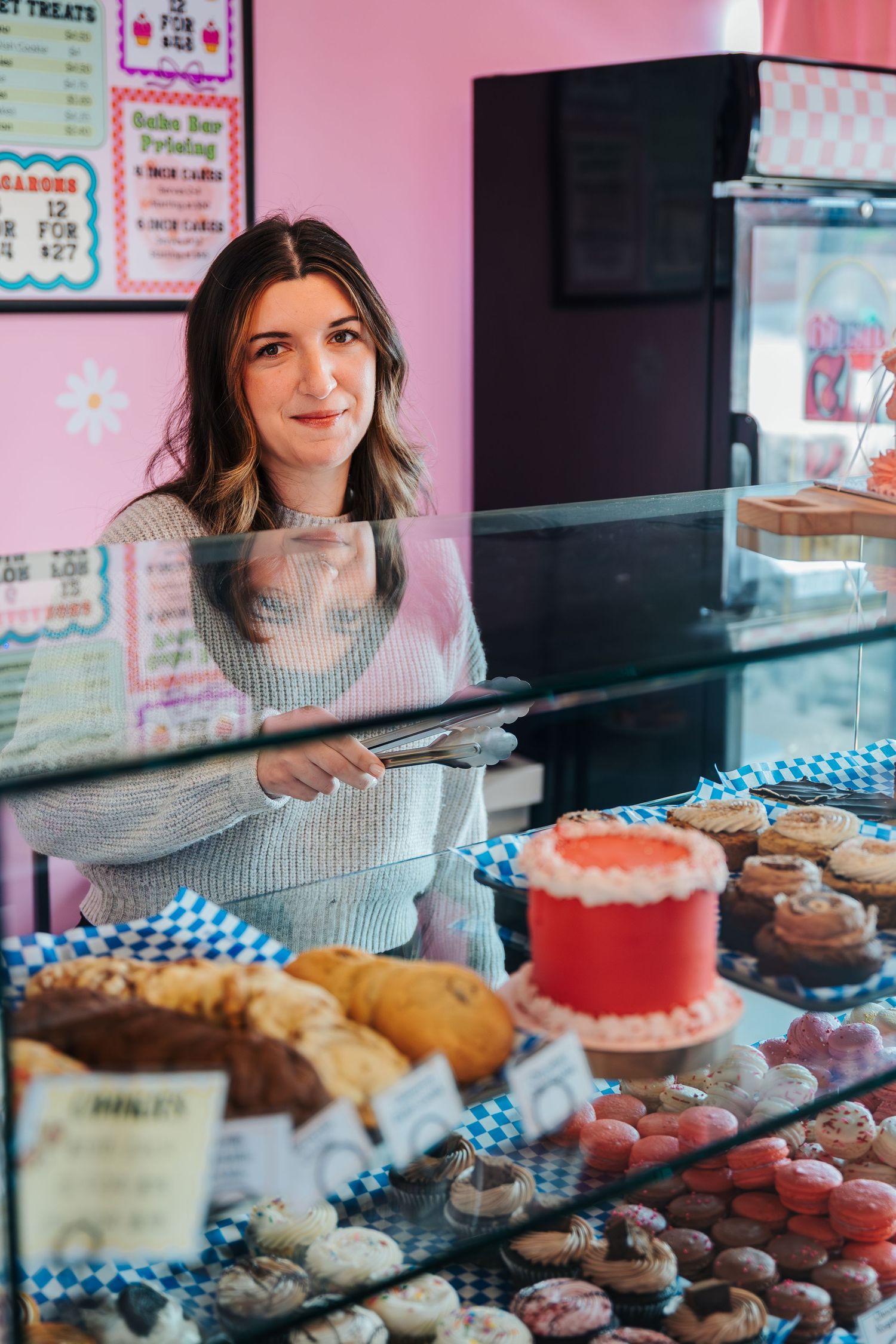 A woman smiles behind a bakery display case filled with colorful cupcakes, donuts, and pastries in a pink shop. Fun and candid photoshoot