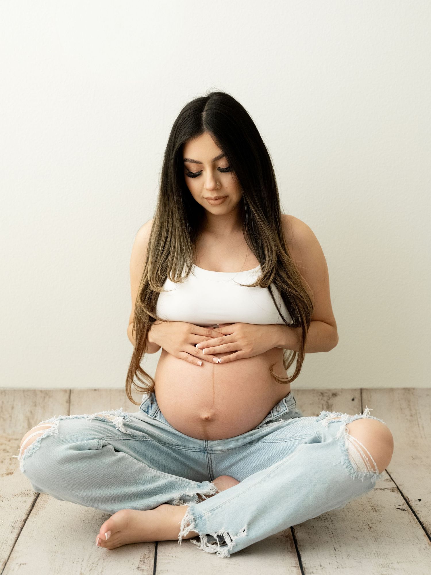 Pregnant woman in white crop top and ripped jeans cradling baby bump against white wall in maternity photo session.