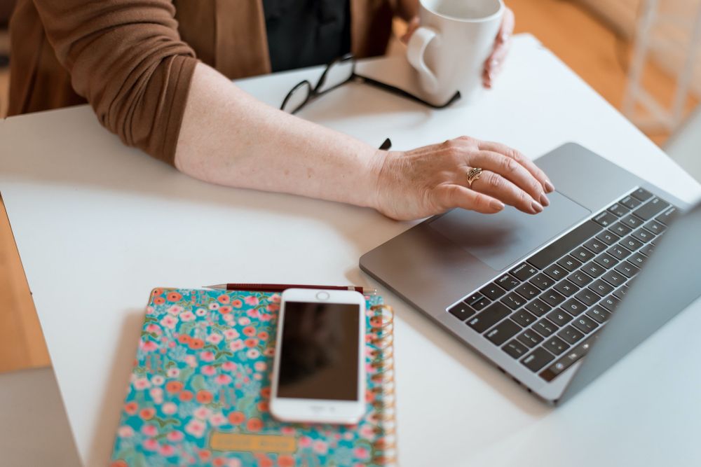 Hand using laptop with floral notebook and smartphone on white desk.