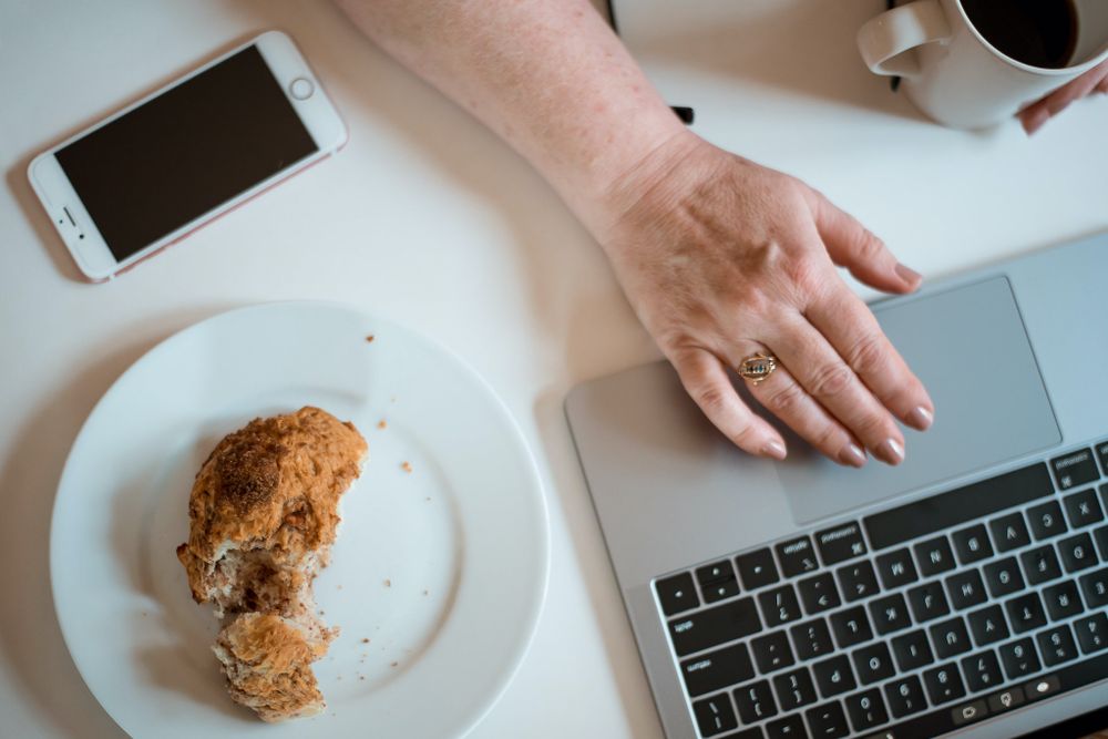 Overhead view of laptop, pastry on plate, and coffee cup on white desk.