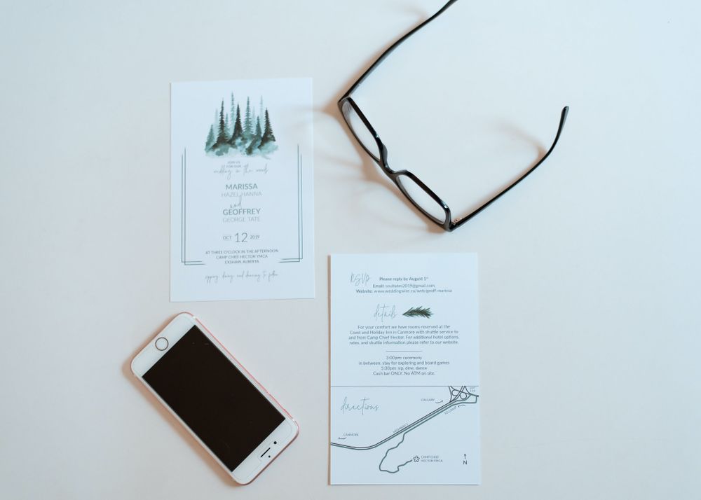 Minimalist wedding invitation with evergreen trees and a phone on white surface.