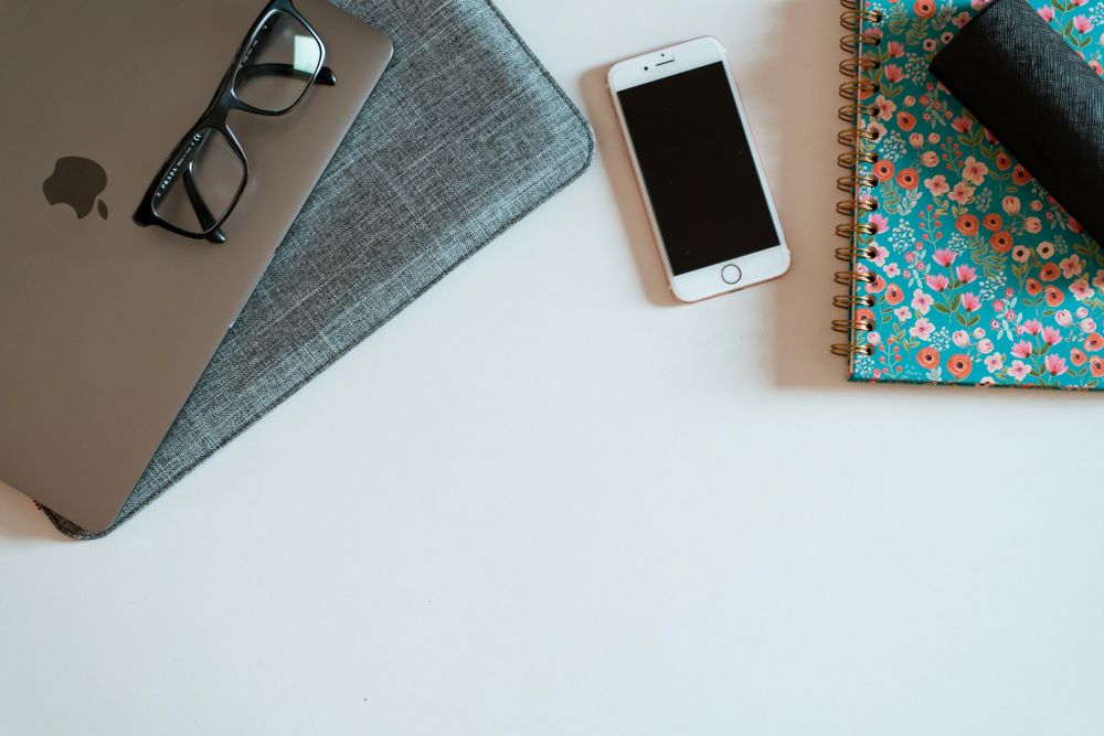 Minimalist desk setup with laptop, glasses, phone and floral notebook on white surface.
