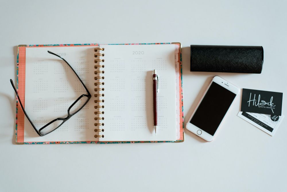 Flatlay of open notebook, glasses, phone and business card on white background.