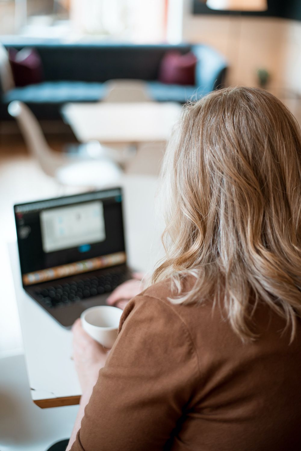 Person in brown sweater working at laptop with coffee cup on desk.