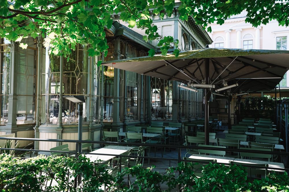 A shaded outdoor cafe terrace with green umbrellas and tables beside a classical colonnade building in Helsinki.
