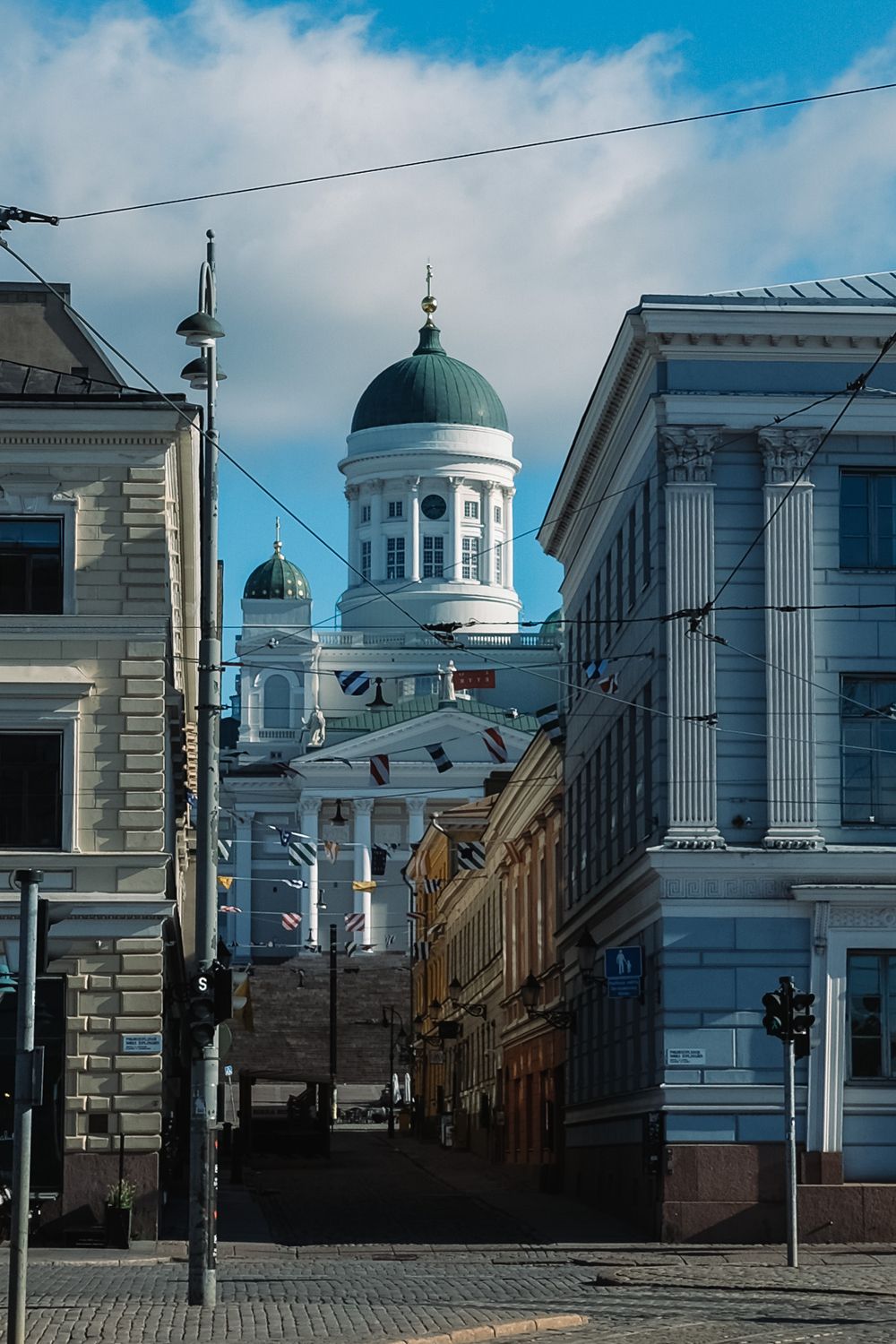 Helsinki Cathedral dome rises above a charming cobblestone street lined with colorful buildings and festive flags.